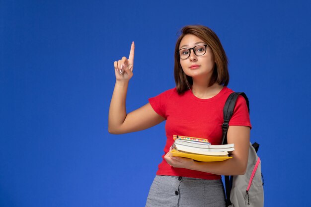 Front view of female student in red shirt with backpack holding books and files on light blue wall