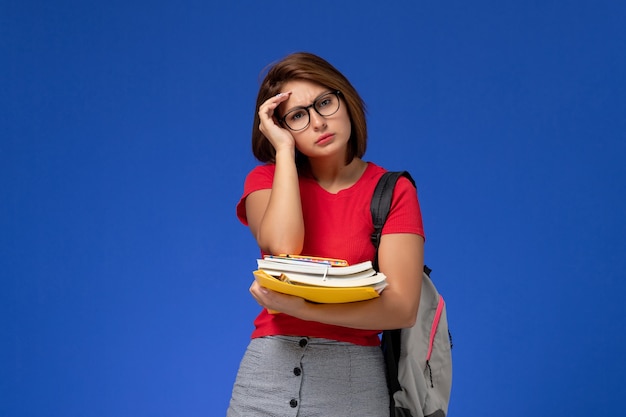 밝은 파란색 벽에 책과 파일을 들고 배낭과 빨간 셔츠에 여성 학생의 전면보기