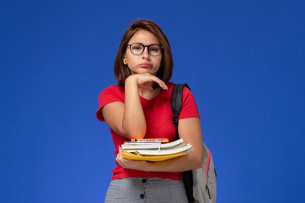 책과 파란색 벽에 생각하는 파일을 들고 배낭과 빨간 셔츠에 여성 학생의 전면보기