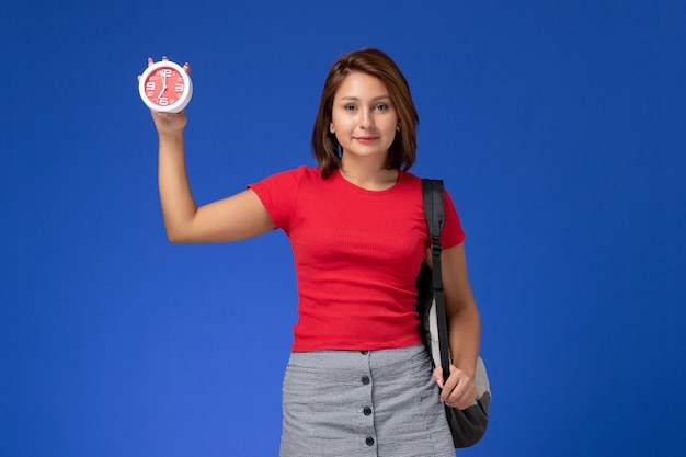 Front view of female student in red shirt with backpack holding clocks on the blue wall