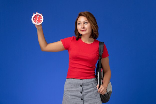Front view of female student in red shirt with backpack holding clocks on the blue wall