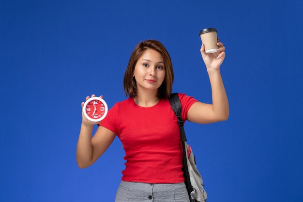 Front view of female student in red shirt with backpack holding clocks and coffee on blue wall