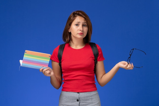 Front view of female student in red shirt with backpack holding copybook on the light-blue wall