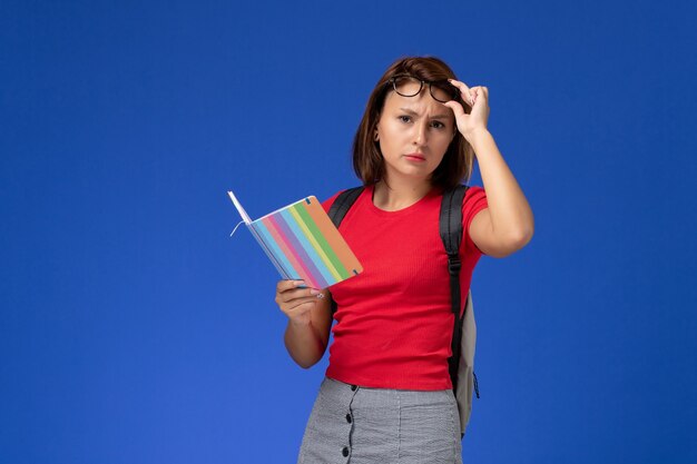 Foto gratuita vista frontale della studentessa in camicia rossa con lo zaino che tiene il quaderno che toglie gli occhiali da sole sulla parete blu