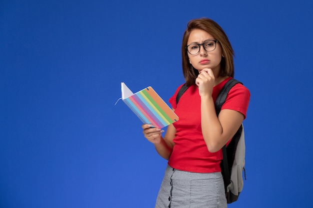 青い壁に考えているコピーブックを保持しているバックパックと赤いシャツの女子学生の正面図