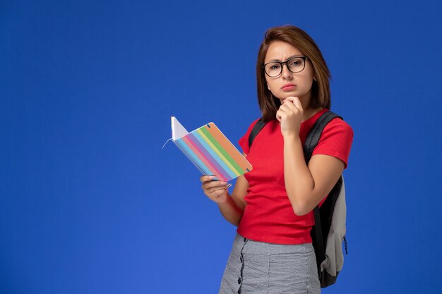 青い壁に考えているコピーブックを保持しているバックパックと赤いシャツの女子学生の正面図