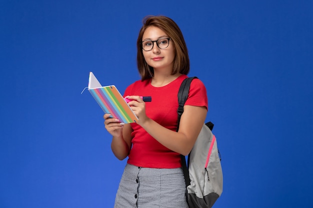 Front view of female student in red shirt with backpack holding felt pens and copybook on blue wall
