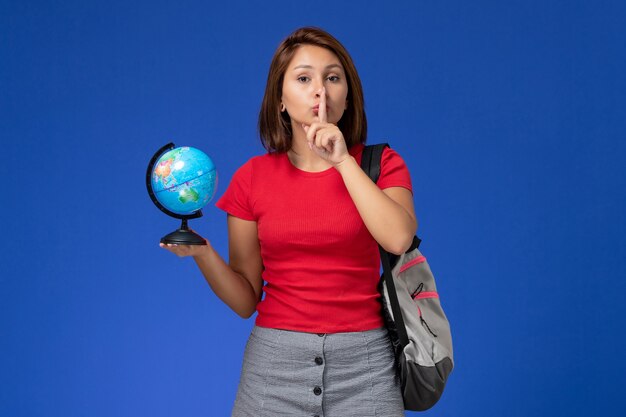 Front view of female student in red shirt with backpack holding little globe on blue wall