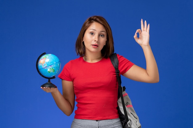 Vista frontale della studentessa in camicia rossa con zaino che tiene piccolo globo sulla parete blu