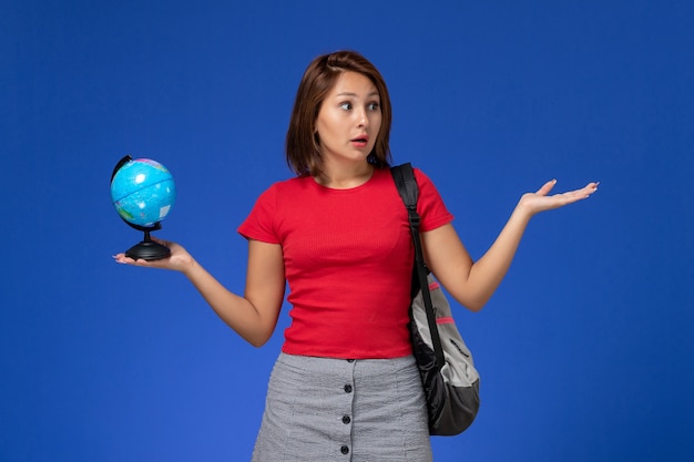 Vista frontale della studentessa in camicia rossa con zaino che tiene piccolo globo sulla parete blu