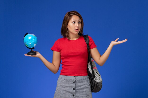 Front view of female student in red shirt with backpack holding little globe on the blue wall