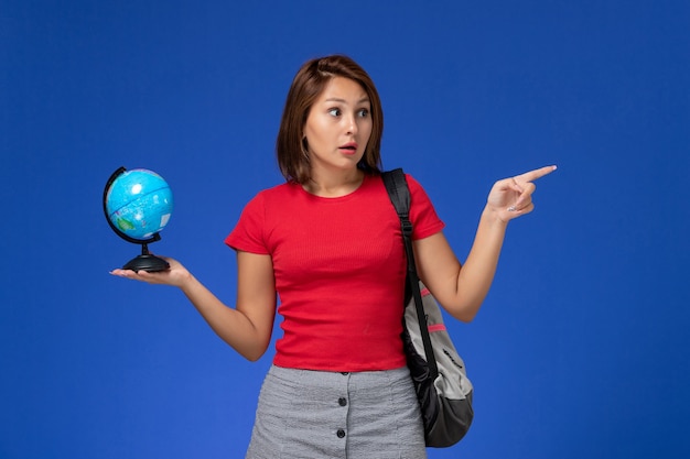 水色の壁に小さな地球儀を保持しているバックパックと赤いシャツの女子学生の正面図