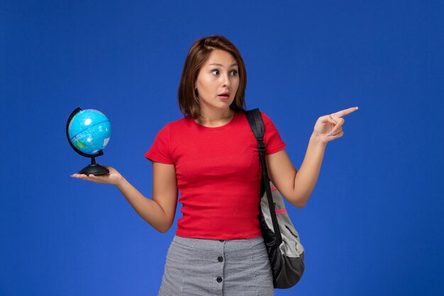 Вид спереди студентки в красной рубашке с рюкзаком, держащей маленький глобус на голубой стене