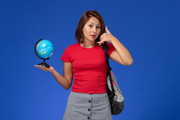 Front view of female student in red shirt with backpack holding little globe posing on blue wall