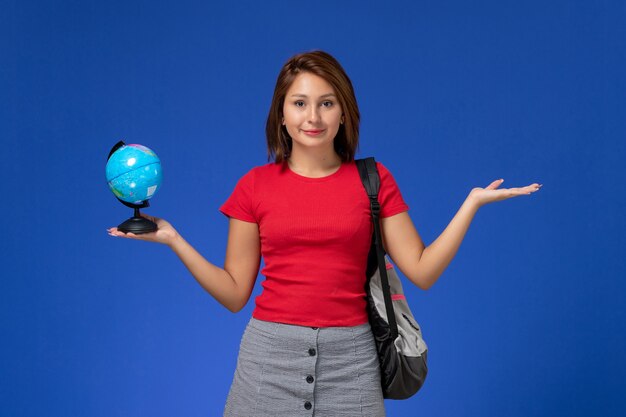 Front view of female student in red shirt with backpack holding little globe and smiling on blue wall