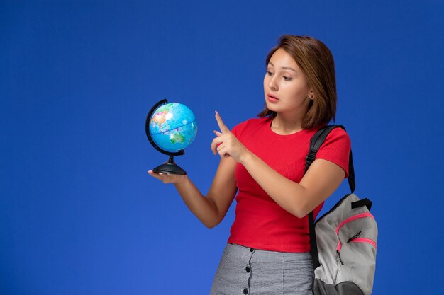 Front view of female student in red shirt with backpack holding little round globe on blue wall