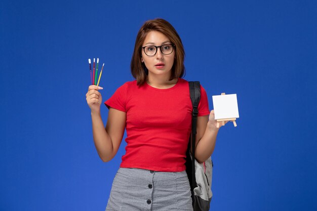 Front view of female student in red shirt with backpack holding tassels for drawing on the light-blue wall