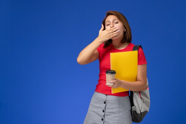 黄色のファイルと青い壁にあくびをしているコーヒーを保持しているバックパックと赤いシャツの女子学生の正面図