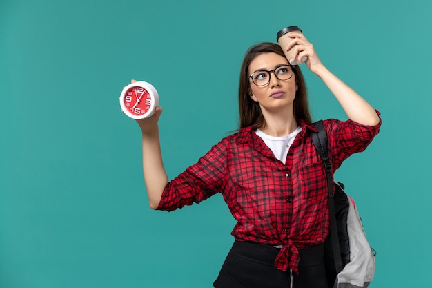 Front view of female student wearing backpack holding clocks and coffee on blue wall