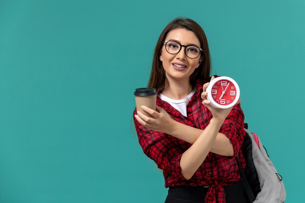 Front view of female student wearing backpack holding clocks and coffee on the blue wall