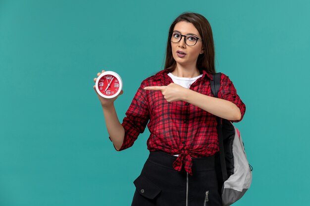 Front view of female student wearing backpack holding clocks on the light blue wall