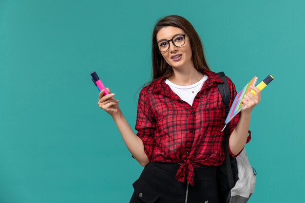 Front view of female student wearing backpack holding copybook and felt pens on blue wall