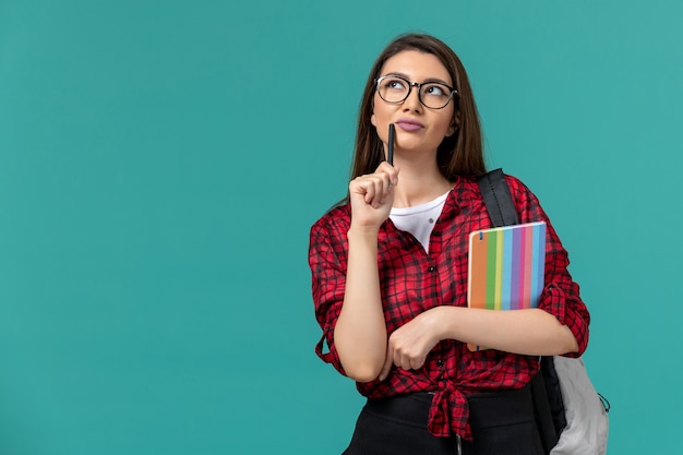 Front view of female student wearing backpack holding copybook and pen thinking on blue wall