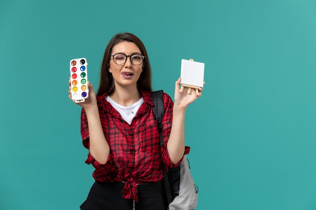 Front view of female student wearing backpack holding easel and paints winking on light-blue wall