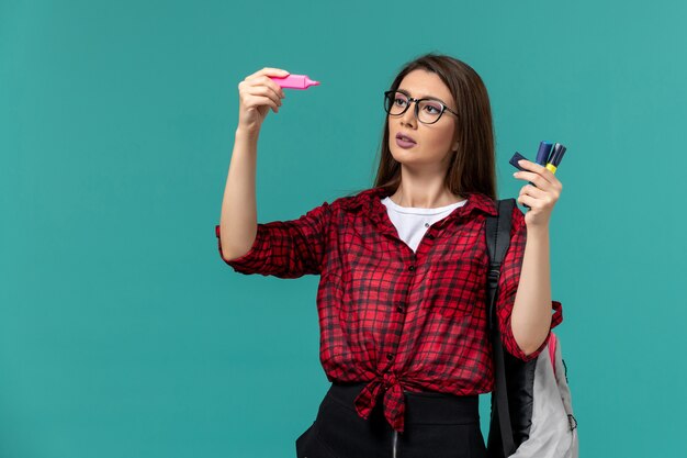 Front view of female student wearing backpack holding felt pens on blue wall