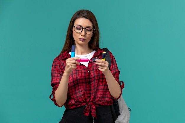 Front view of female student wearing backpack holding felt pens on blue wall