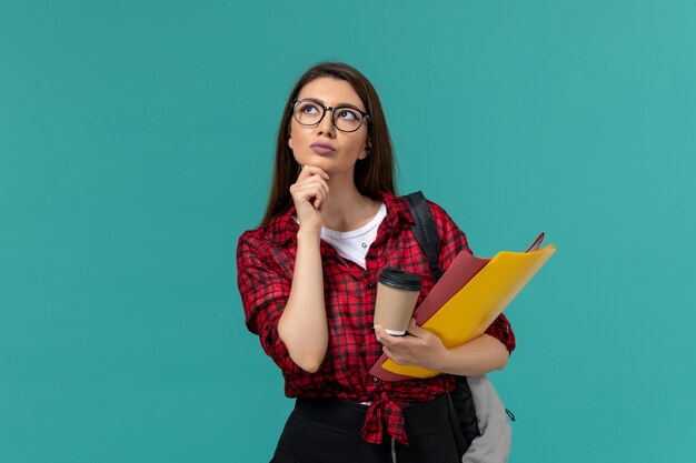 Front view of female student wearing backpack holding files and coffee thinking on light-blue wall