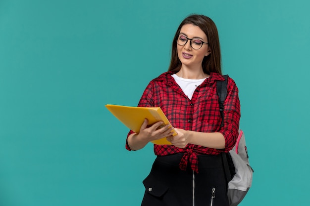 Front view of female student wearing backpack and holding files on the light blue wall