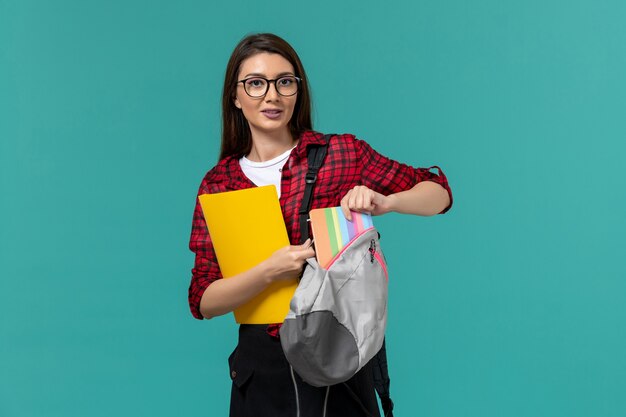 Front view of female student wearing backpack and holding files taking out copybook on the light-blue wall