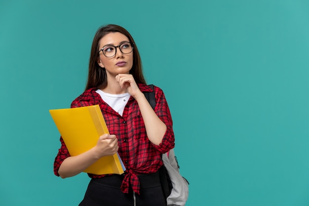 Front view of female student wearing backpack and holding files thinking on light-blue wall