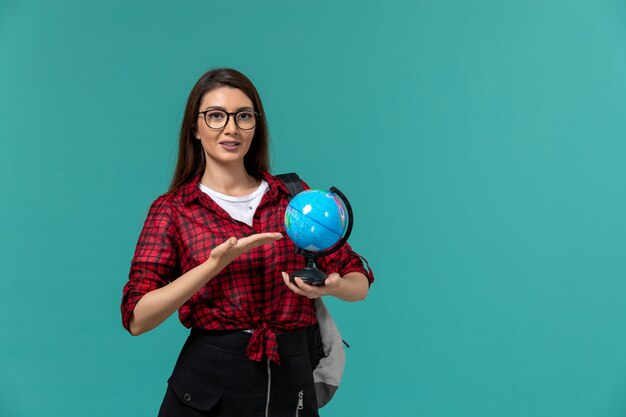 Front view of female student wearing backpack holding little globe on light-blue wall