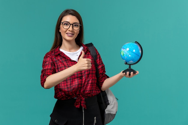 Front view of female student wearing backpack holding little globe on light blue wall