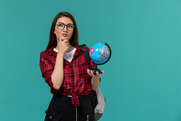 Front view of female student wearing backpack holding little globe on light blue wall