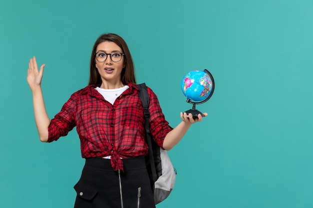 Front view of female student wearing backpack holding little globe on light blue wall