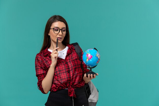 Front view of female student wearing backpack holding little globe and pen on light blue wall