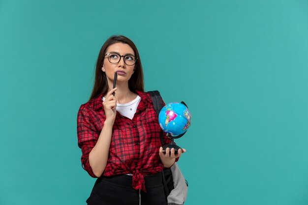 Front view of female student wearing backpack holding little globe and pen on light blue wall