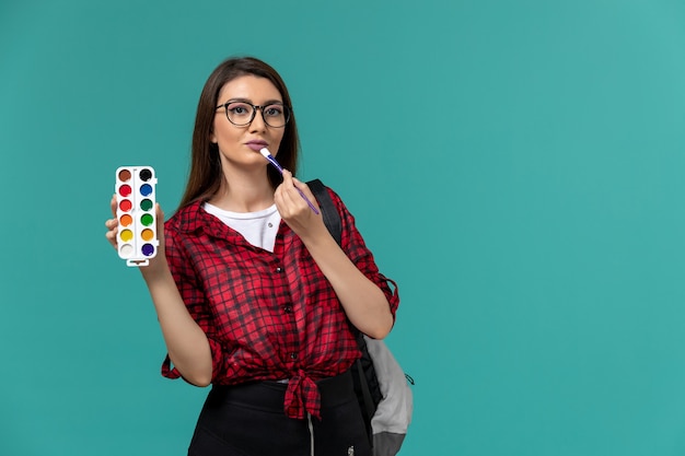 Front view of female student wearing backpack holding paints and tassels on blue wall