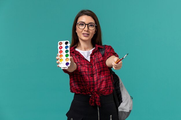 Front view of female student wearing backpack holding paints and tassels on blue wall