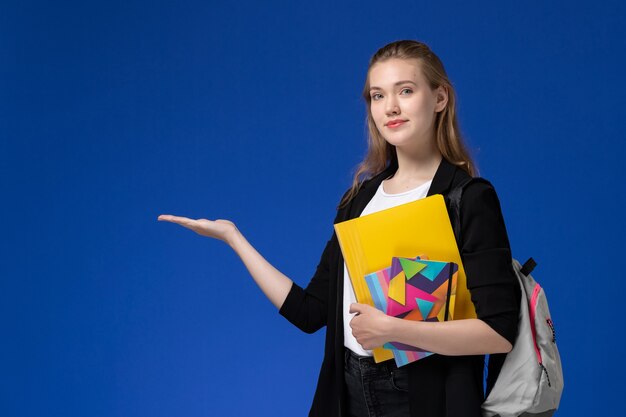 Front view female student in white shirt and black jacket wearing backpack holding files with copybooks on blue wall college university lessons