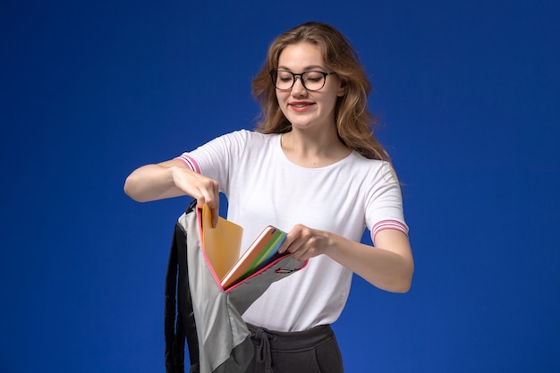 Free photo front view of female student in white shirt holding backpack and yellow files on the blue wall