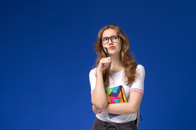 Front view of female student in white shirt holding pen and copybook thinking on the blue wall