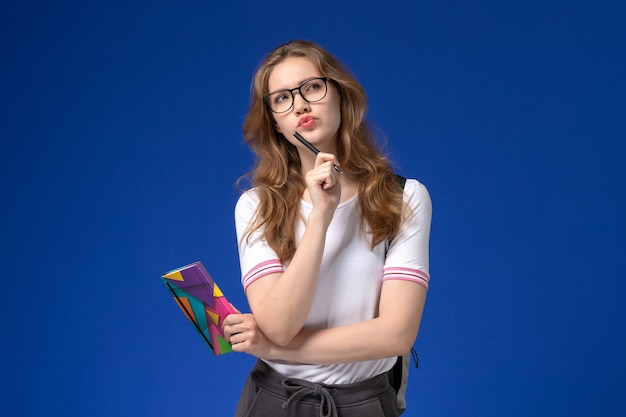 Front view of female student in white shirt holding pen and copybook thinking on the blue wall