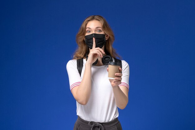 Front view of female student in white shirt wearing backpack black mask and holding coffee on the blue wall