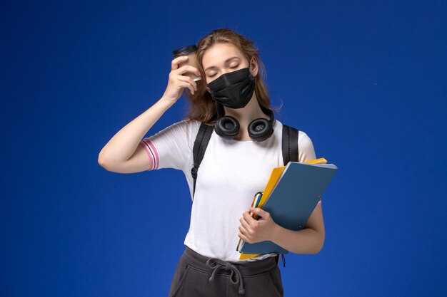 Front view of female student in white shirt wearing backpack black mask holding coffee and files on the blue wall