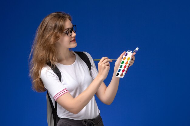 Front view of female student in white shirt wearing backpack and holding art paint brushes on the blue wall