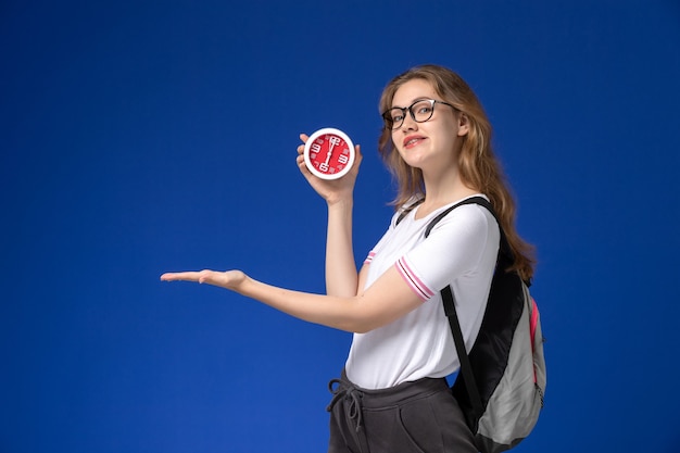 Vista frontale della studentessa in camicia bianca che indossa zaino e che tiene gli orologi sulla femmina blu del college di lezione dell'università dello scrittorio